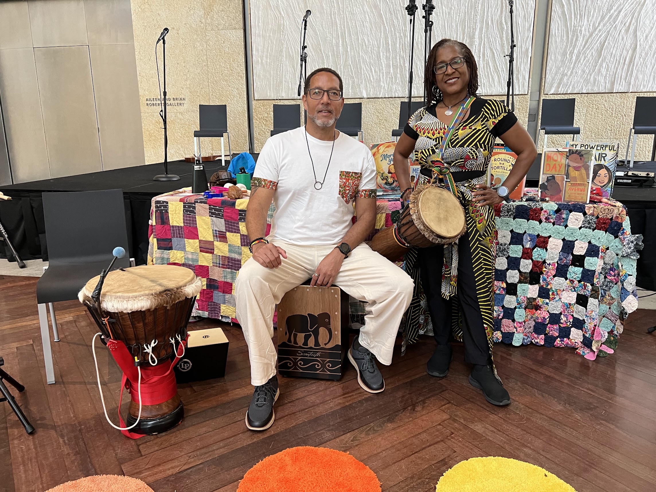 Leslie holding a drum beside a seated musician, with quilts and children’s books