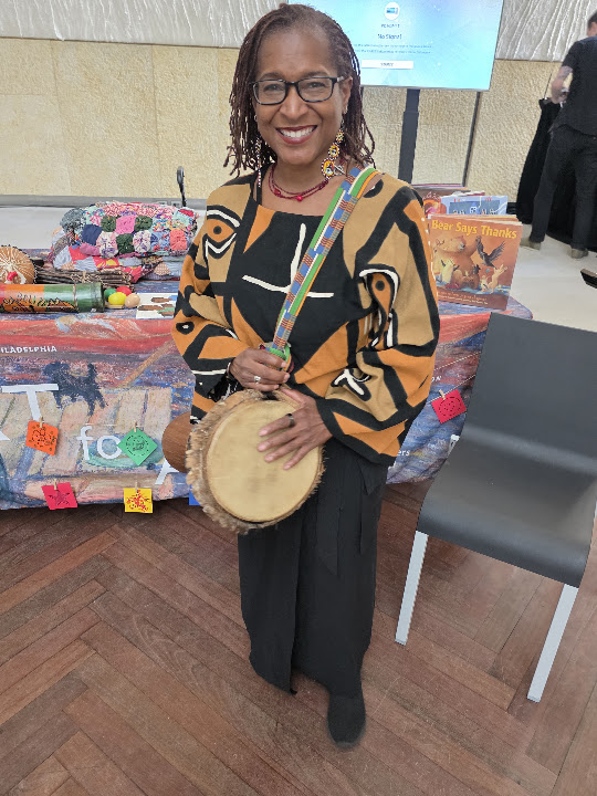 Leslie smiling and holding a drum, wearing a black and gold patterned top